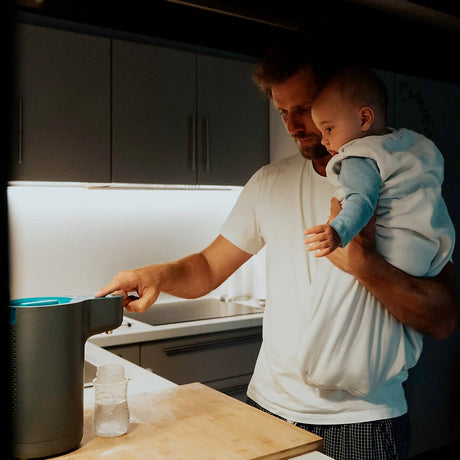 Vater mit Baby auf dem Arm, steht in der Nacht in der Küche an einem Babyflaschen Automat 