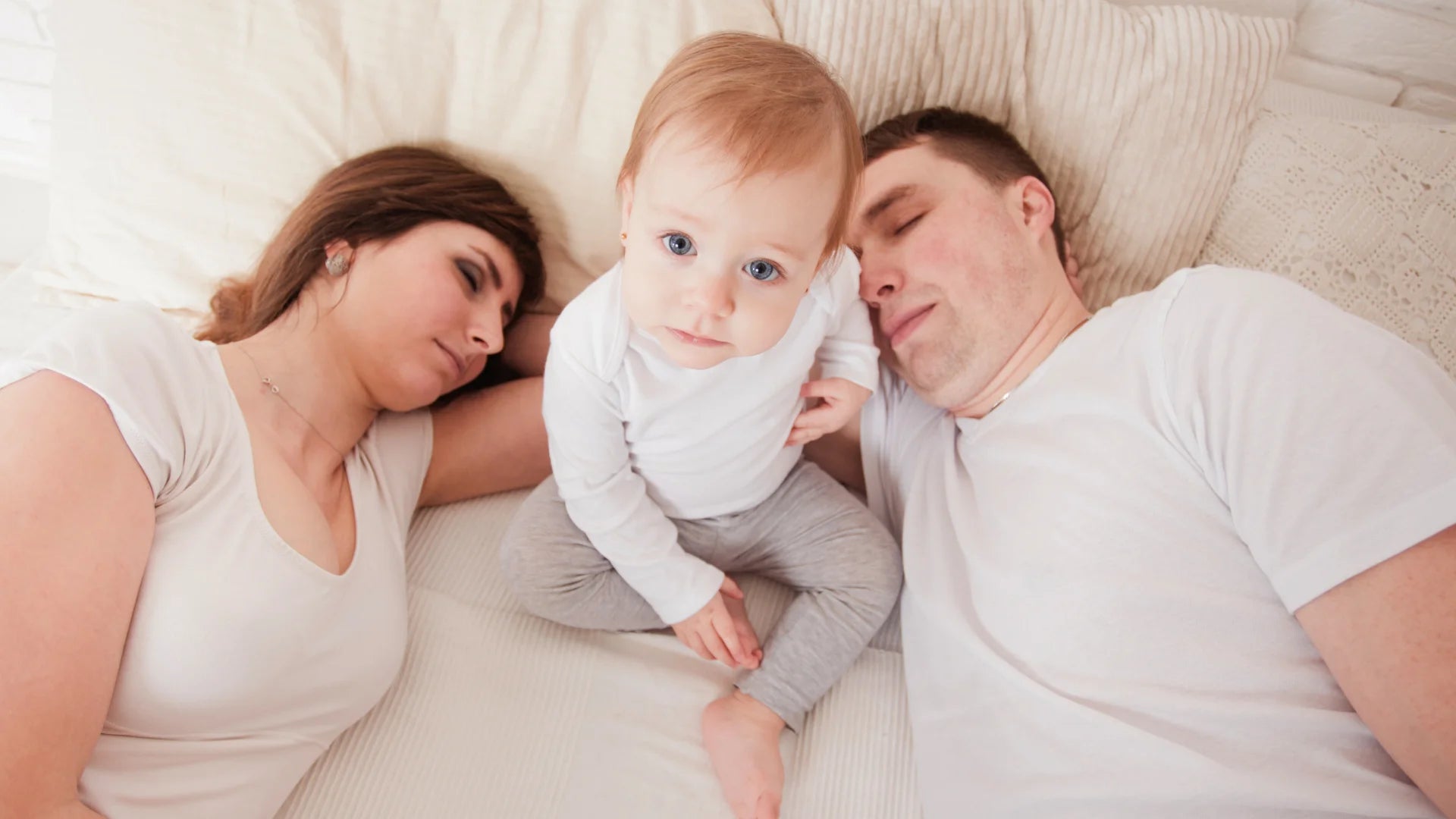 Eltern liegen im Bett und versuchen zu schlafen, Baby sitzt zwischen ihnen und scheint hellwach zu sein