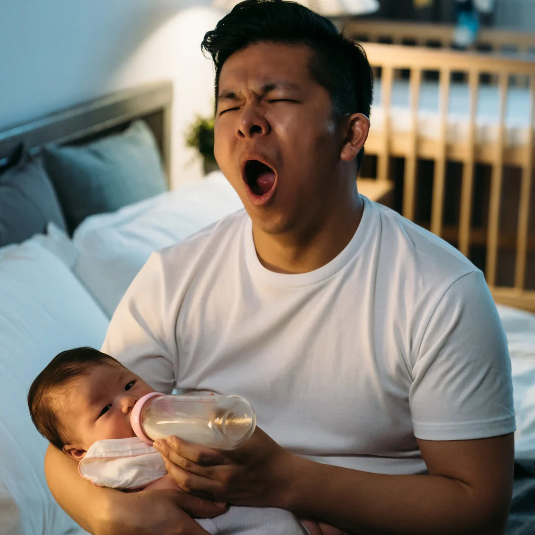Vater sitzt mitten in der Nacht auf der Bettkante und füttert sein Baby mit der Flasche, dabei gähnt er, weil er müde ist