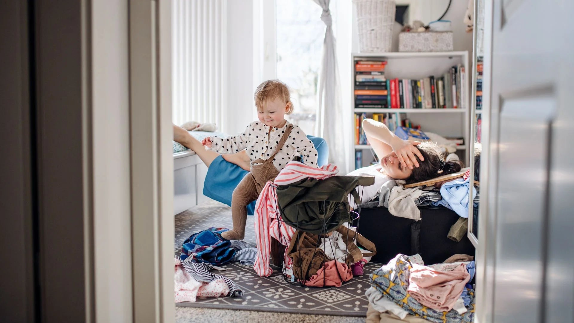 Eine Mutter versinkt im Chaos, also Wäschebergen, und ihr Kleinkind läuft glücklich durch die Gegend